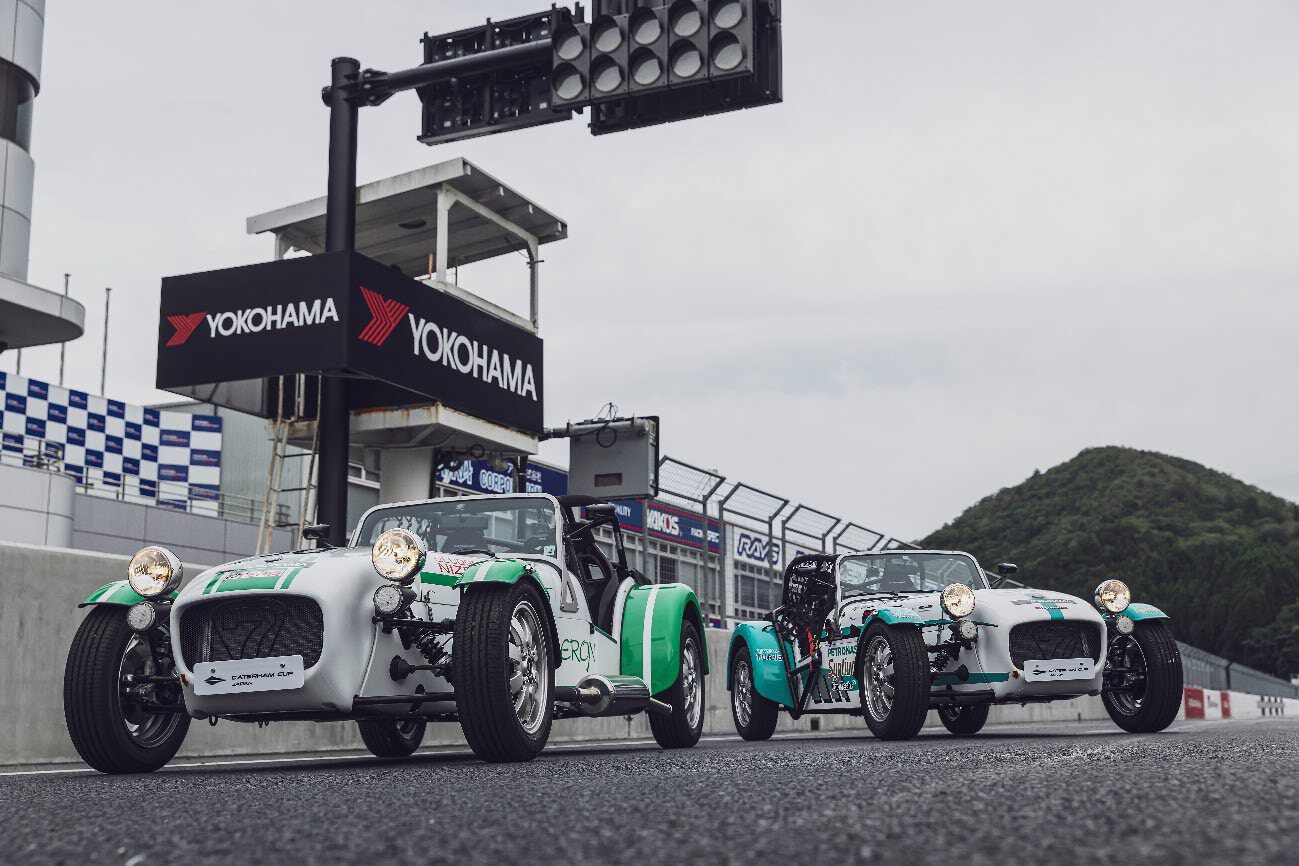 Two vintage open-top race cars sit on a racetrack beneath a Yokohama sign, with pit wall and a hillside in the background.